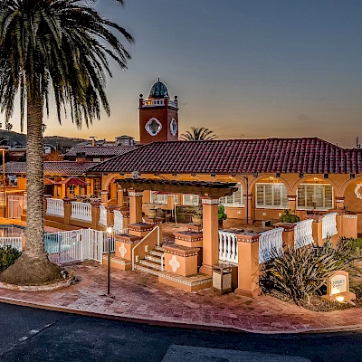 The image shows a Spanish-style building with a clock tower, surrounded by palm trees and featuring a pool area. The scene is illuminated at dusk.