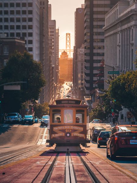 A streetcar travels uphill in an urban setting with tall buildings on either side, and the sun setting in the distance, creating a warm glow.