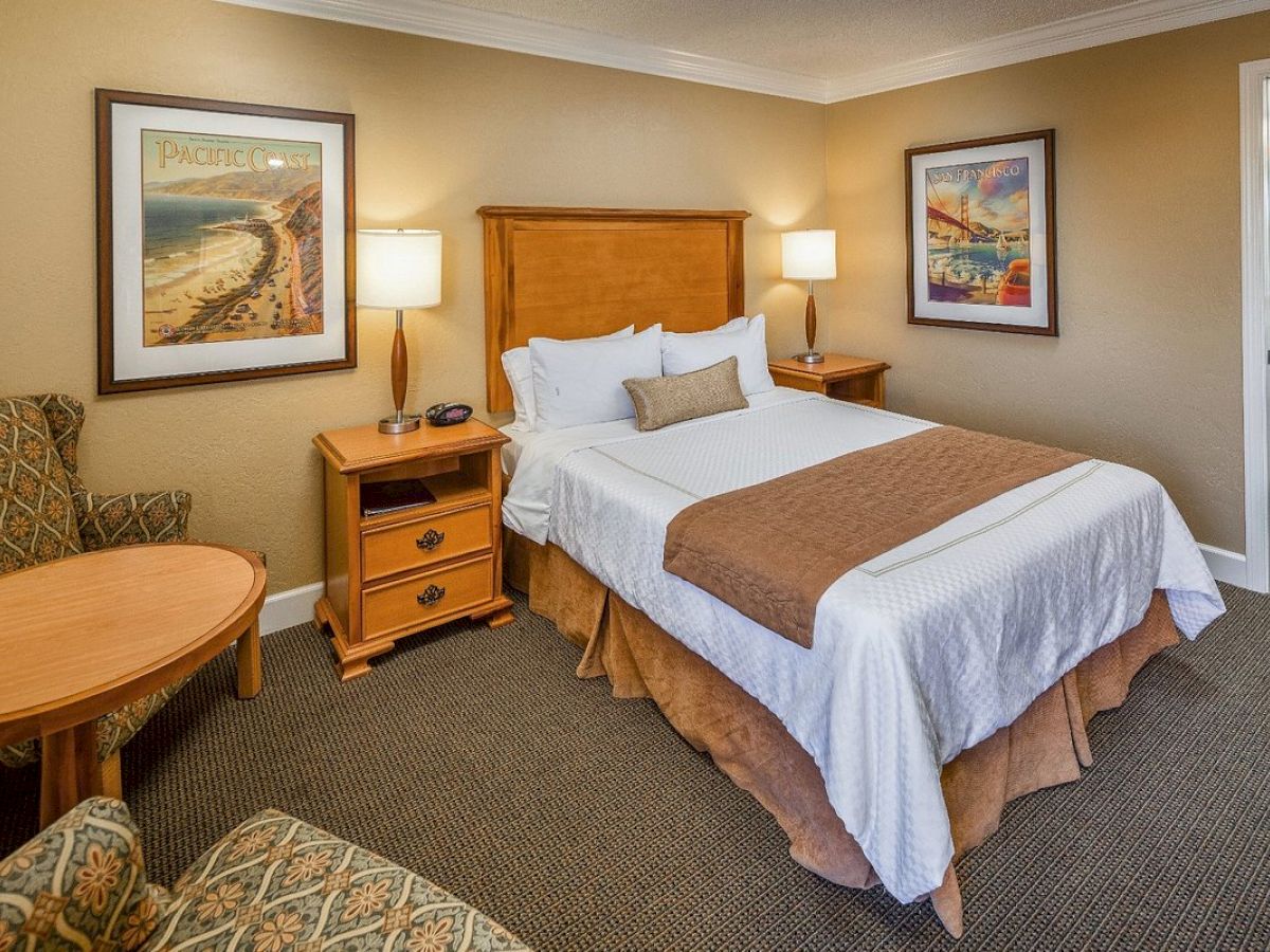 A cozy hotel room with a single bed, nightstands, armchairs, a round table, framed art, and a bathroom entrance.