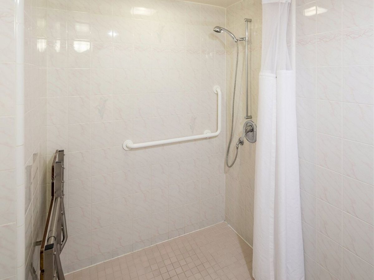 The image depicts a bathroom shower area equipped with a wall-mounted showerhead, a grab bar, and a foldable shower seat, with a white shower curtain.