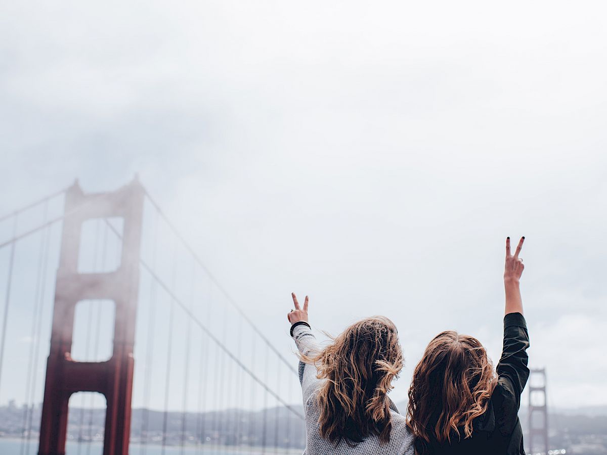 Two people with raised hands making peace signs in front of the Golden Gate Bridge on a cloudy day.