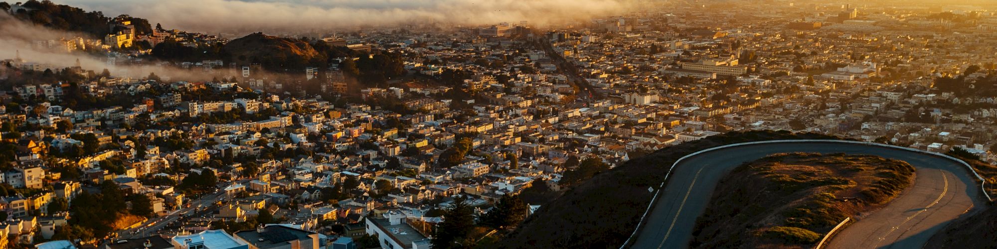 A panoramic view of a city with buildings, fog covering parts of the skyline, and a winding road leading down from a hill at sunrise or sunset.