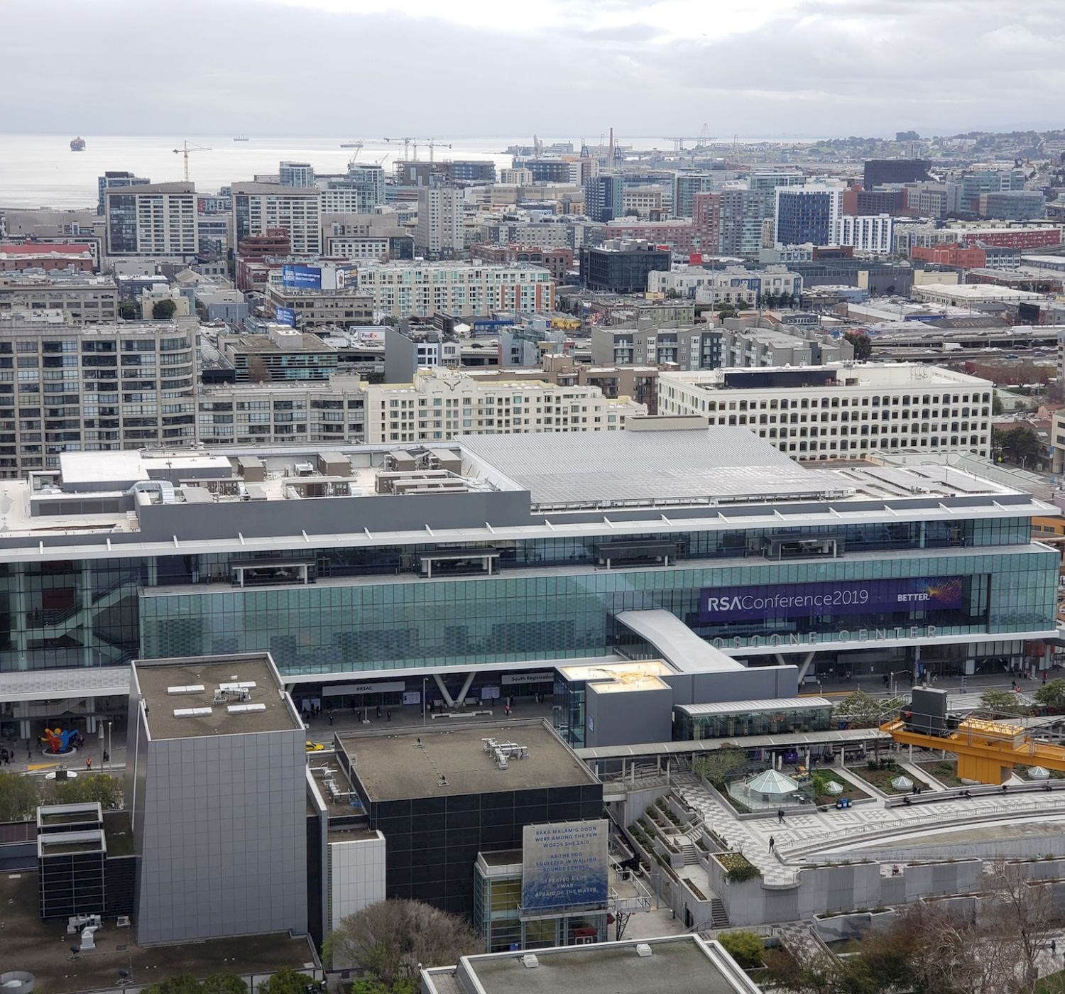 This aerial view shows a large convention center building in a bustling city with numerous surrounding buildings and a distant waterfront.