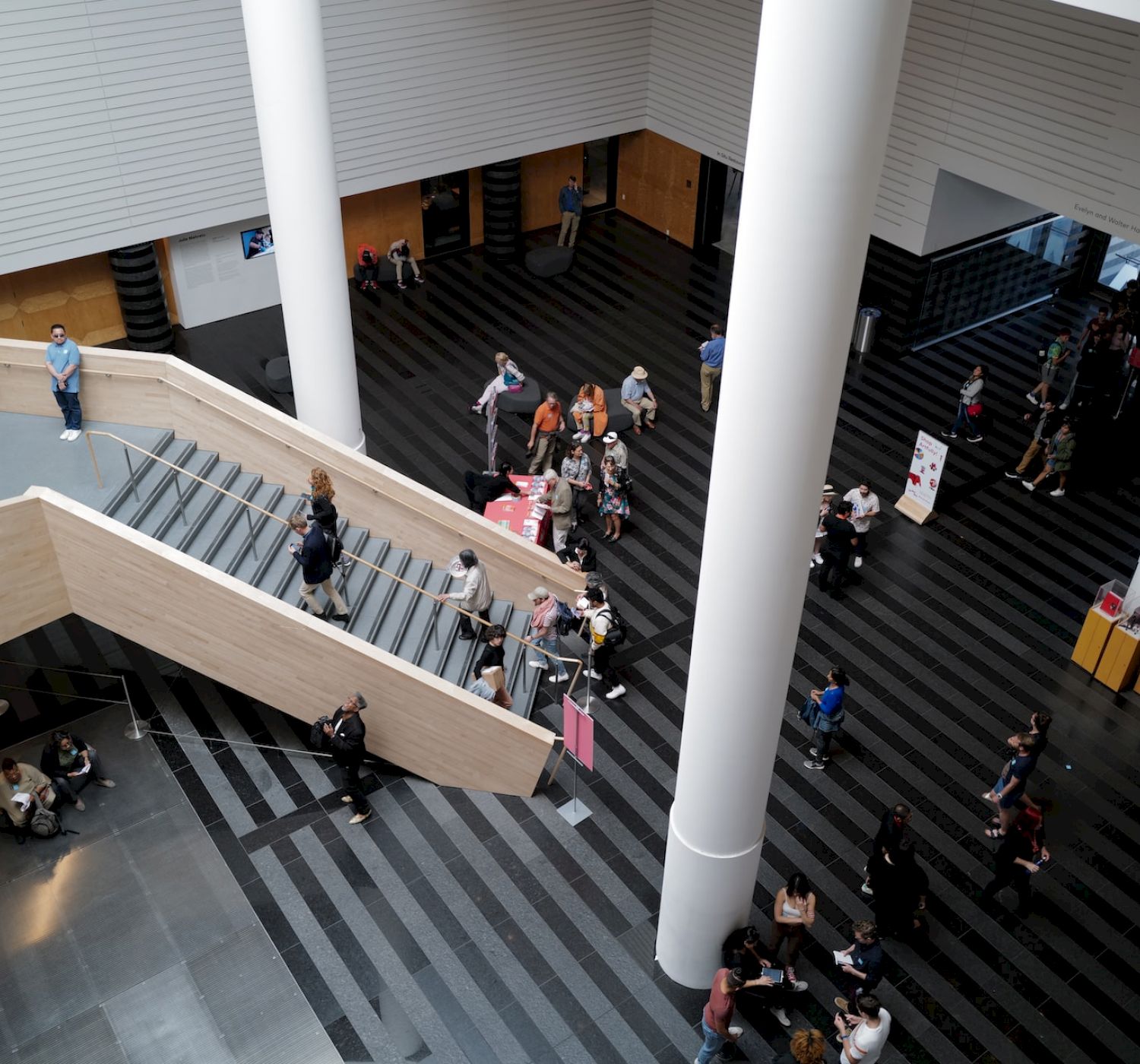 People walking in a spacious indoor area with tall columns, a staircase, and striped flooring, some sitting, others moving around.