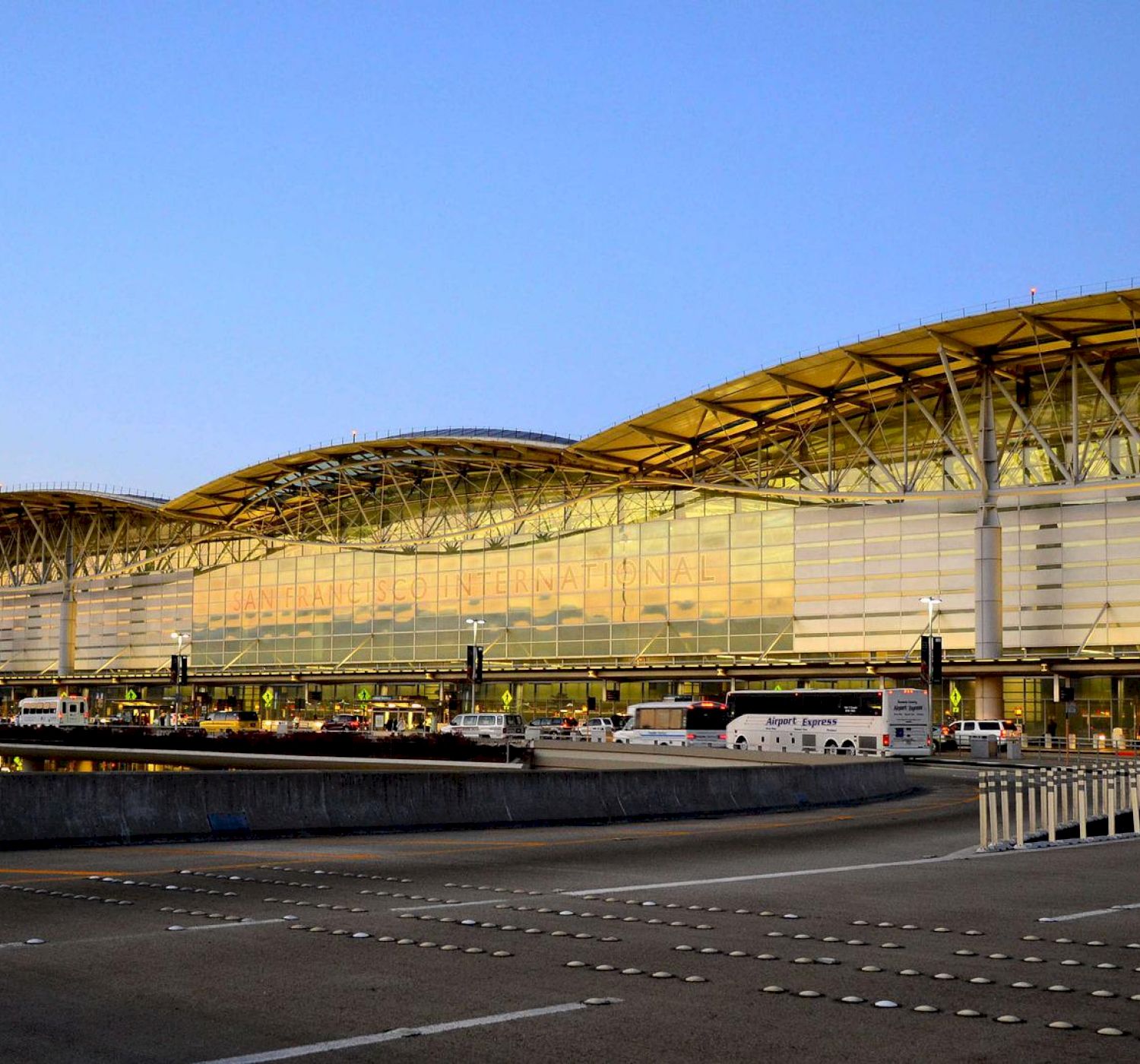 The image shows a modern airport terminal with a wavy roof, large windows, and a roadway in the foreground. There are vehicles and people near the entrance.