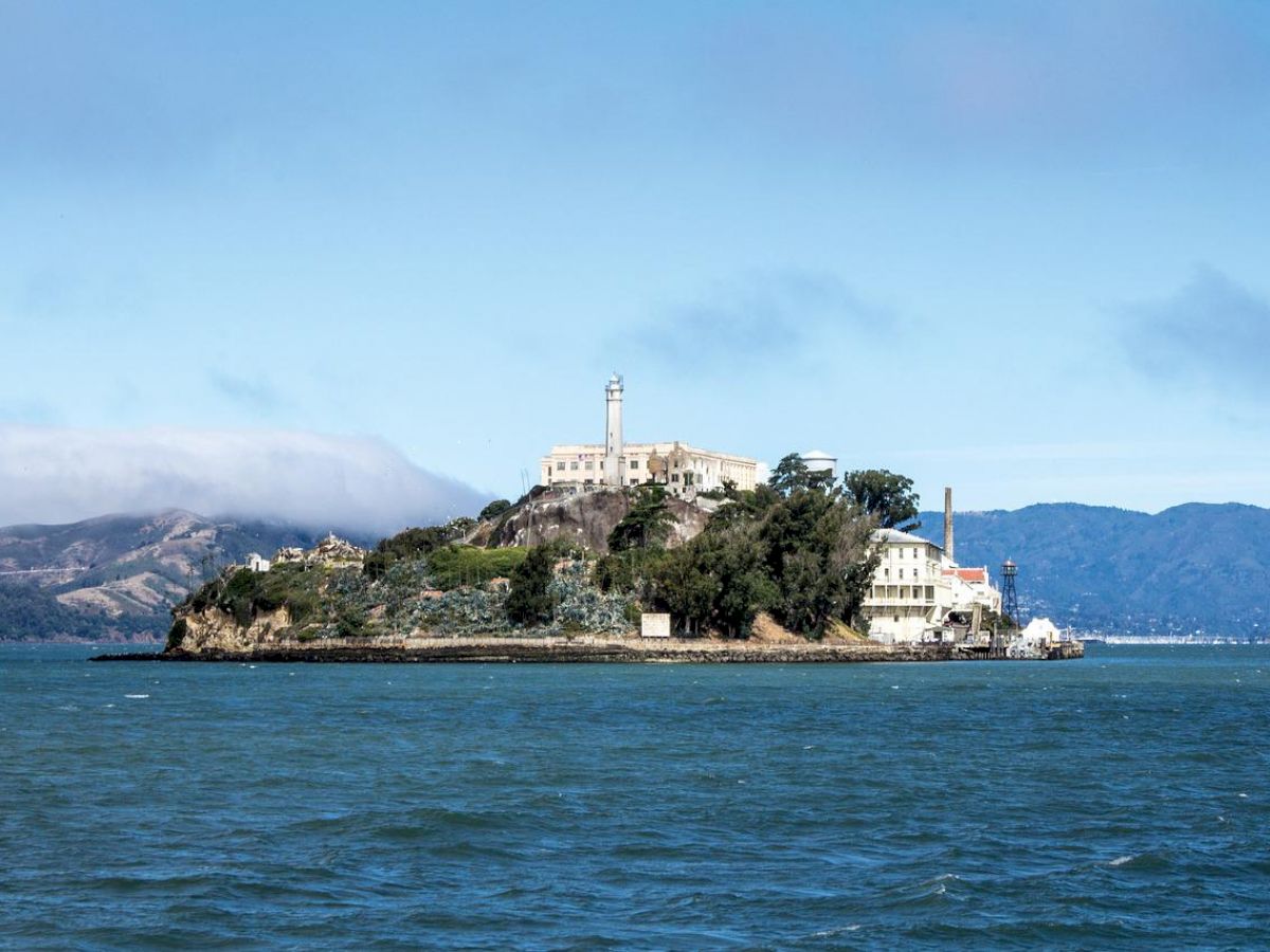 An island with a large historic building surrounded by a body of water, with mountains visible in the background under a partly cloudy sky.