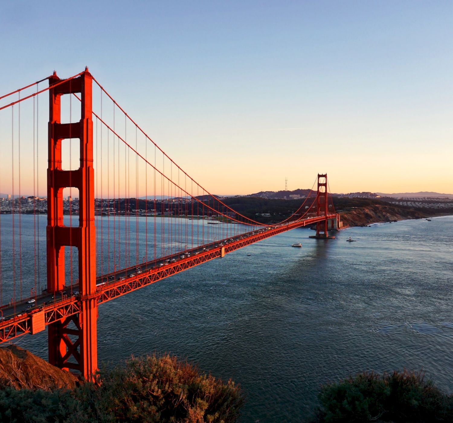This image shows the Golden Gate Bridge at sunset, with the reddish-orange structure spanning across a body of water.