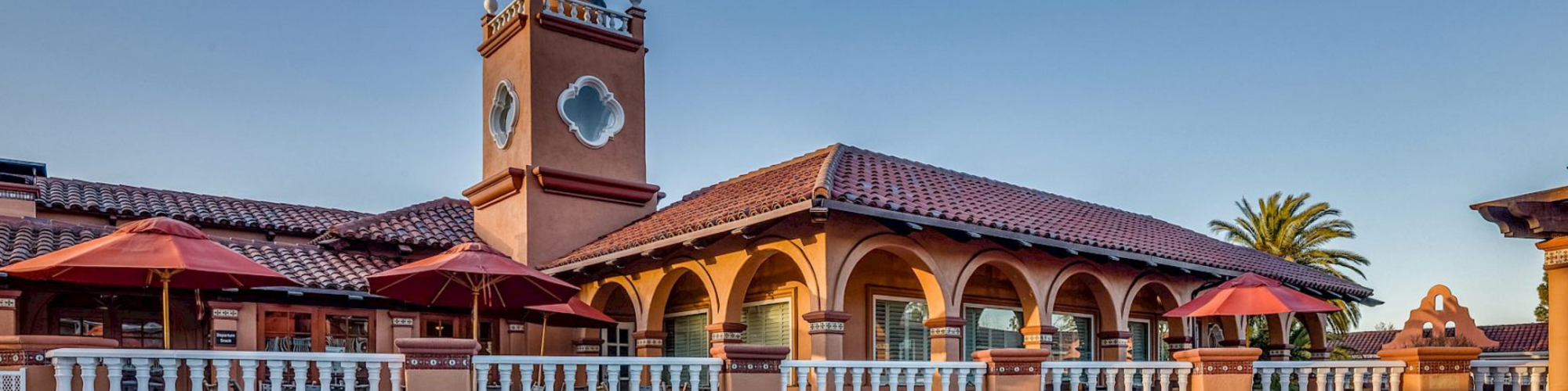 The image features a building with Spanish-style architecture, red-tiled roofs, arched windows, a tower, and a patio area with red umbrellas.